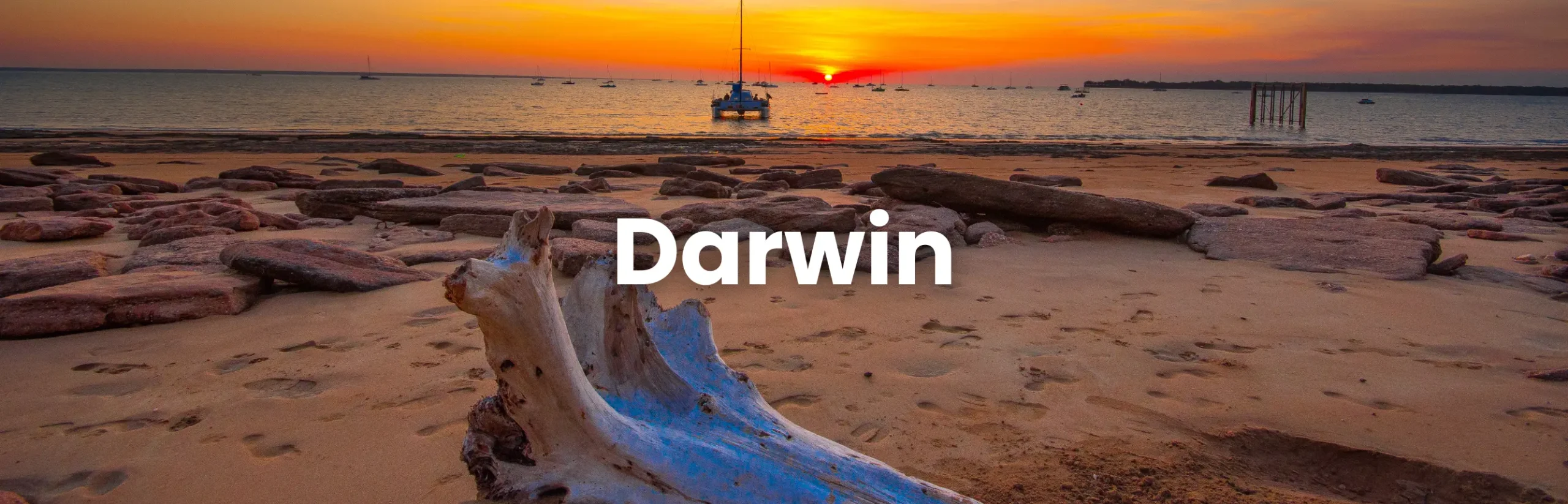 Darwin Conference location featuring a scenic beach at sunset with driftwood, rocks and anchored boats on the horizon.
